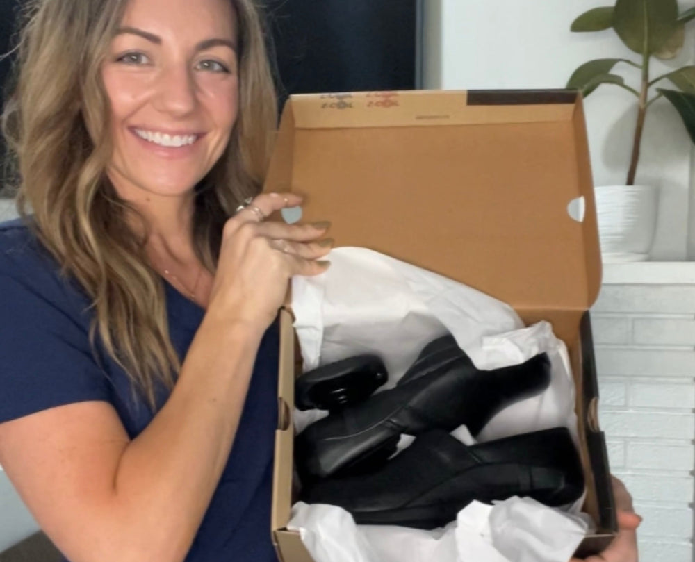 A woman in navy blue scrubs holds an open cardboard shoe box lined with white tissue paper, revealing a pair of black ankle boots inside. The setting is a home interior with a white brick fireplace, a television, and a potted plant in the background.